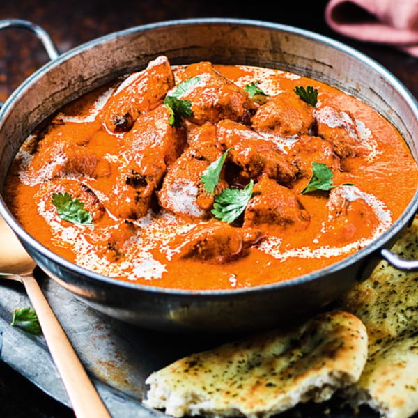 A metal pot filled with creamy orange Indian butter chicken, garnished with cilantro, next to naan bread.