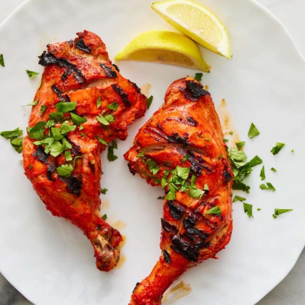 Two grilled Indian chicken drumsticks, garnished with fresh cilantro and lemon wedges on a white plate.