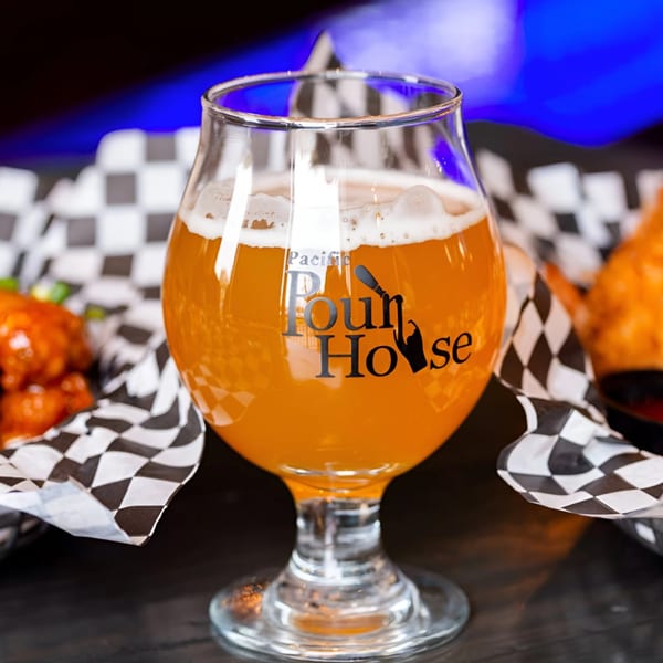 A Pacific PourHouse branded glass of amber beer with foam, next to checkered paper-lined baskets of food.